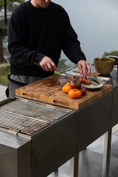 A man in a black sweatshirt grills food on a stainless steel outdoor barbecue grill next to a wooden cutting board with tomatoes and onions.